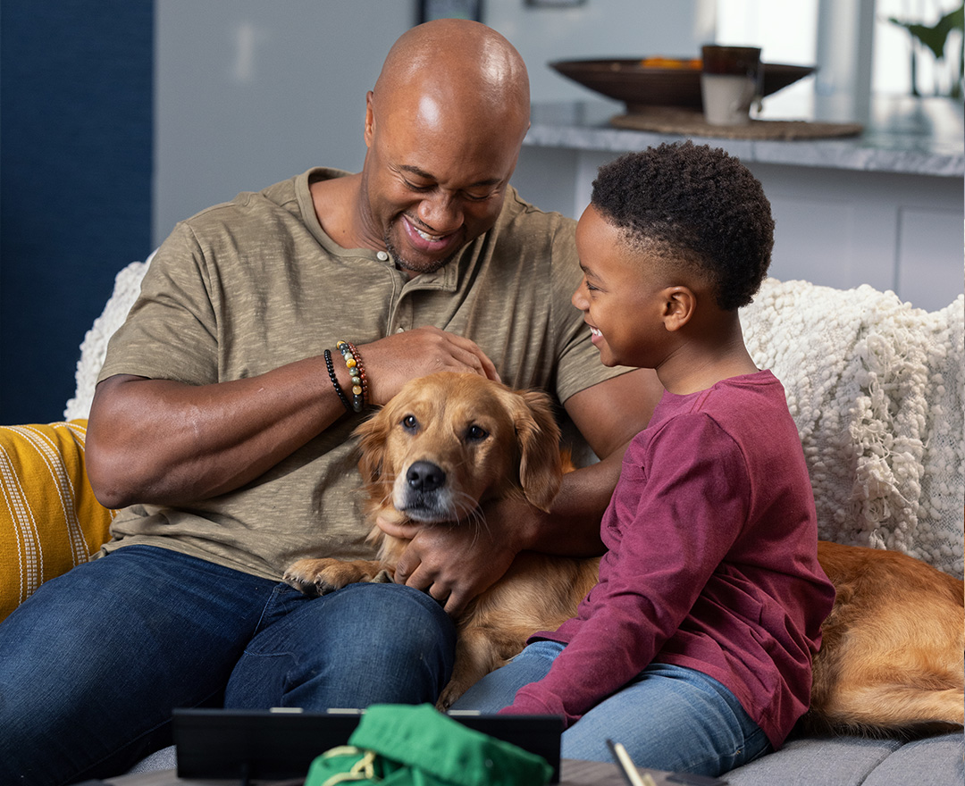 he man seen at the top of the webpage is now sitting comfortably on a couch, smiling with his son and petting his dog.